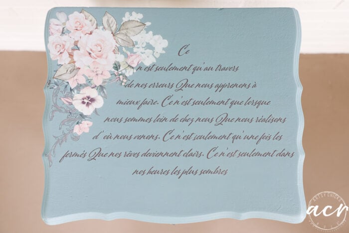A blue plaque, perfect for an end table, features painted white and pink flowers in the top left corner and French cursive text in black across the center; ACY logo in white is at the bottom right.