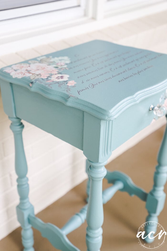 A small, vintage-style end table painted light blue with elegant, turned legs. The tabletop features hand-painted pink and white flowers and scripted text, creating a decorative, shabby-chic look with artistic paint details.