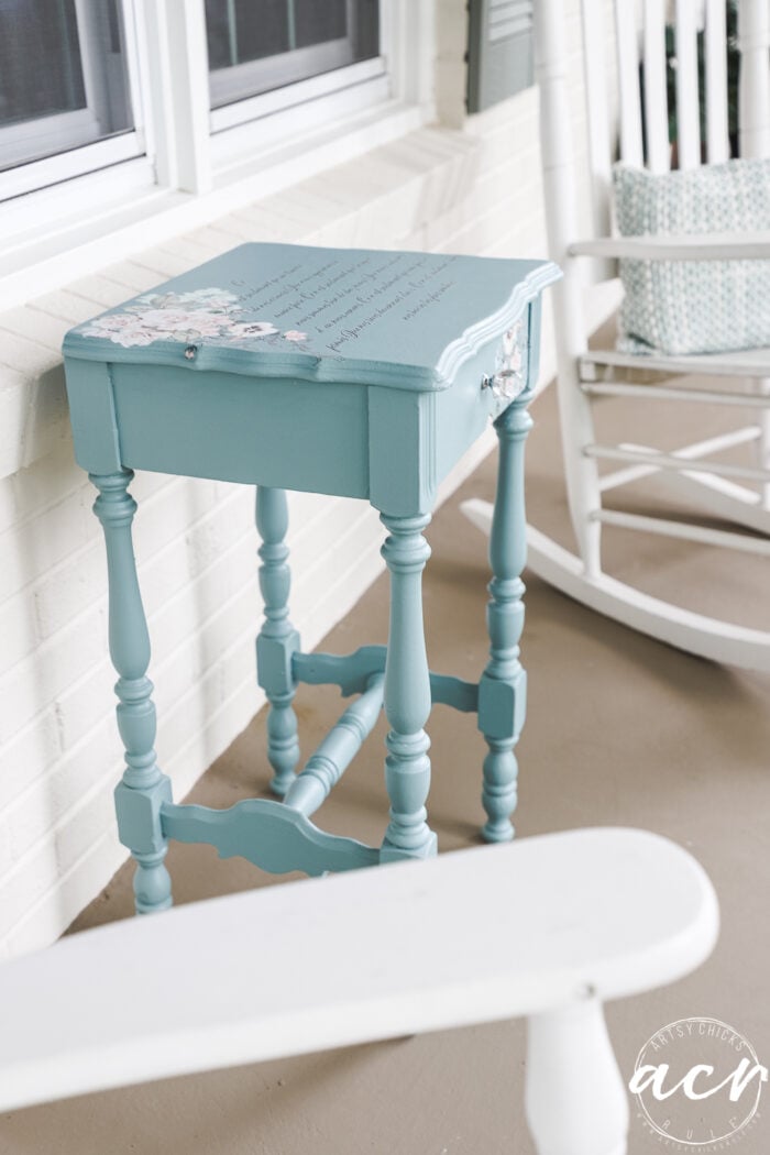 A small, vintage blue end table with floral accents and cursive writing created using a transfer technique sits on a porch next to a white rocking chair and cushion. The porch features a light brown floor and classic white brick walls.