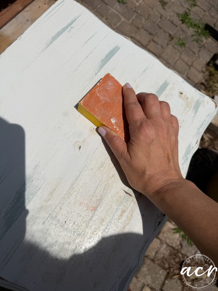 A hand holds a sanding block, smoothing the surface of a white, distressed end table outdoors on a sunny day. The table shows signs of wear, and paving stones are visible in the background.