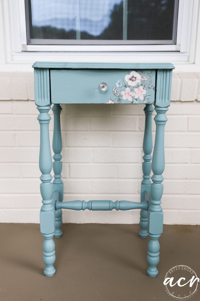 A small vintage end table painted teal with decorative turned legs, featuring a floral transfer design and a clear knob on the single drawer, placed in front of a white brick wall and window.
