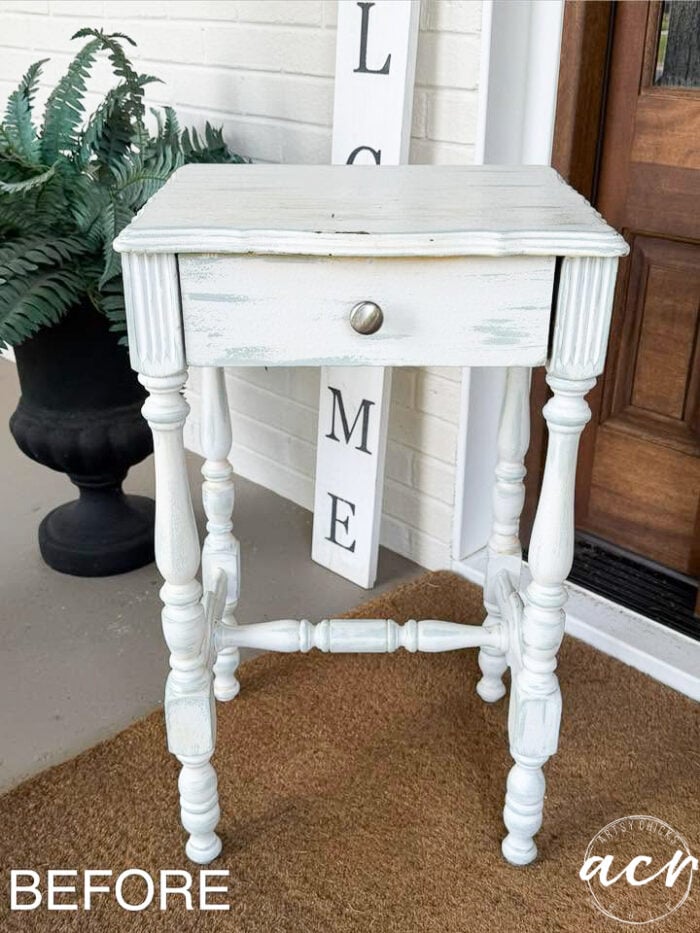 A small, distressed white painted end table with one drawer stands on a brown mat by a front door. A potted fern and a vertical WELCOME sign are in the background. The word BEFORE is in the bottom left corner.