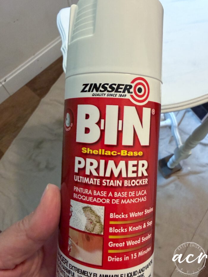 A hand holds a can of Zinsser B-I-N Shellac-Base Primer, highlighting its ultimate stain-blocking power for paint projects. The label notes it blocks water stains, knots, sap, and seals wood in 15 minutes. An end table is visible in the background.