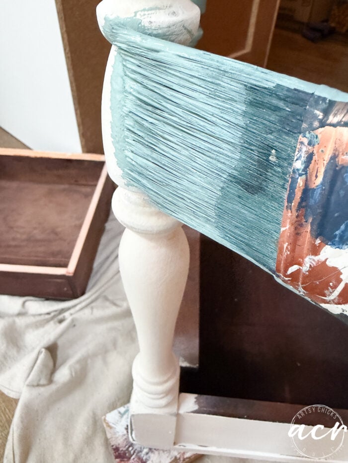 A large paintbrush applies teal paint to the leg of a white wooden end table. A transfer cloth and a wooden drawer sit on the floor in the background.