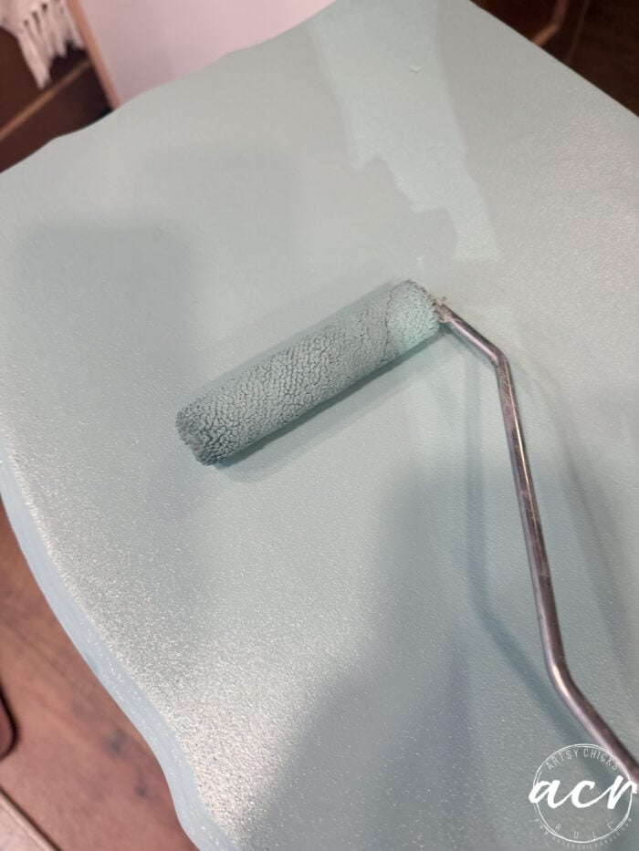 A close-up of a paint roller applying light blue paint to a smooth end table, likely transferring color evenly. The background reveals a hint of wooden flooring and a white object.