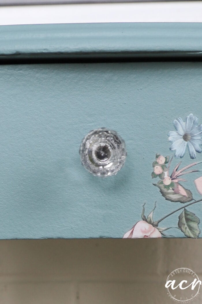 A close-up of a blue painted end table drawer with a clear glass knob and a floral transfer design featuring pink and blue flowers on the front.