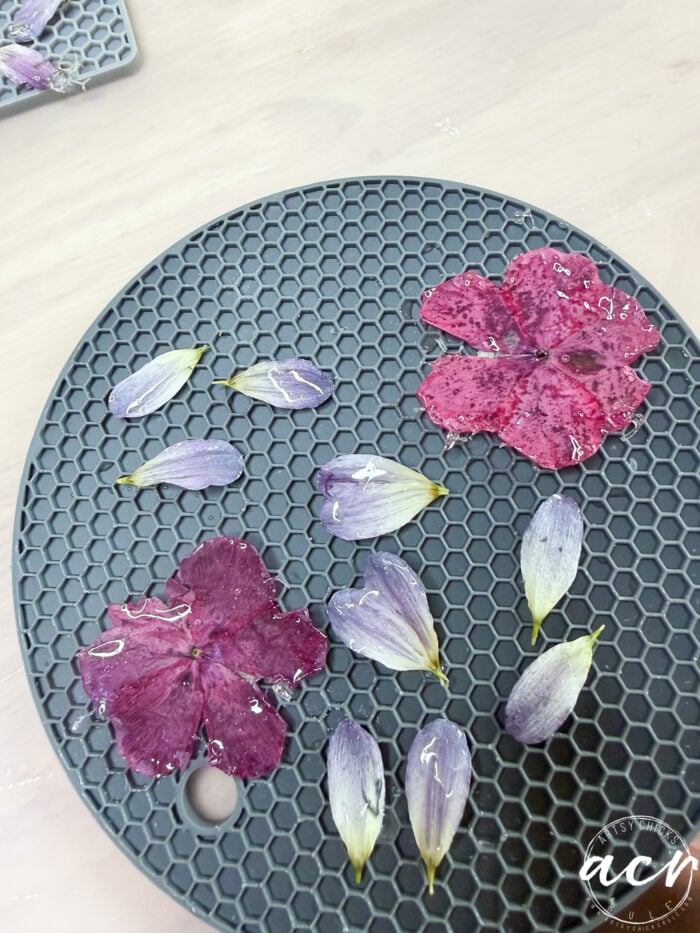 Pressed purple and magenta flower petals are arranged on a round, black, honeycomb-patterned mat, evoking the delicate beauty of flower petal resin earrings. The surface appears slightly wet, with a white logo in the bottom right corner.