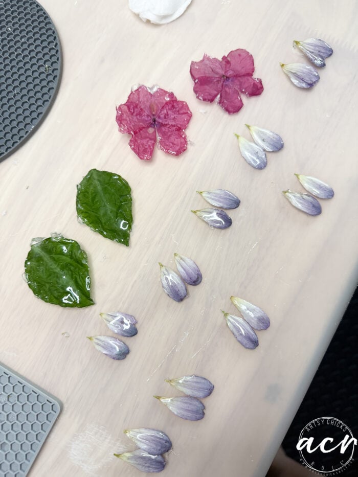 Pressed pink flowers, green leaves, and purple petal buds are arranged in neat rows on a light surface&mdash;perfect inspiration for DIY earrings or resin earrings. A round gray mat and a white logo in the lower right corner are also visible.