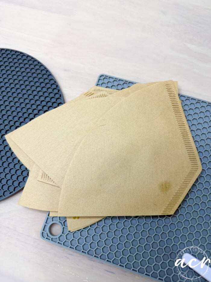 Three brown paper coffee filters are stacked on a gray silicone mat with a textured pattern, placed on a light-colored surface. Nearby, delicate flower petal resin earrings add an artistic touch to the serene scene. A dark placemat is partially visible in the background.