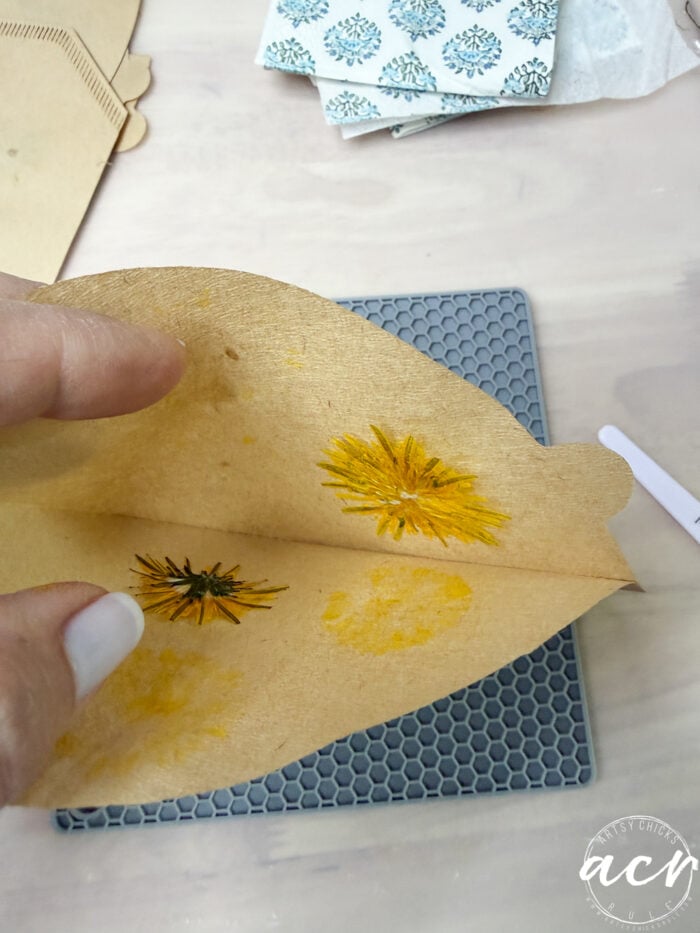 A hand holds an open brown envelope with two yellow pressed flowers inside, over a textured gray mat; patterned paper and crafting materials&mdash;perfect for learning how to make flower petal earrings or resin earrings&mdash;are visible in the background.