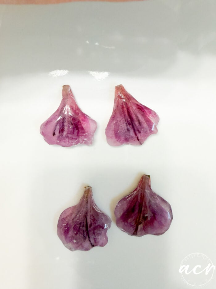 Four translucent, purple, flower-petal resin earrings are arranged on a white surface, with two at the top and two at the bottom. The earrings have a glossy texture and visible vein-like lines.