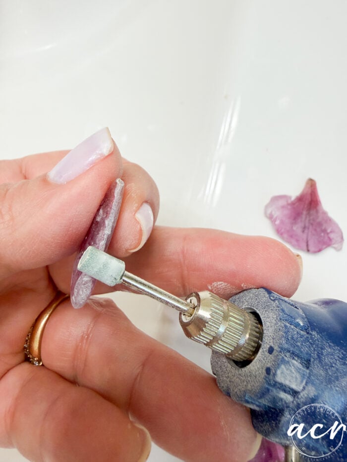 A hand holds a purple petal, using a rotary tool to drill a hole&mdash;an essential step to make resin earrings. Another petal and a white surface are visible. The person has light pink nails and wears a gold ring.