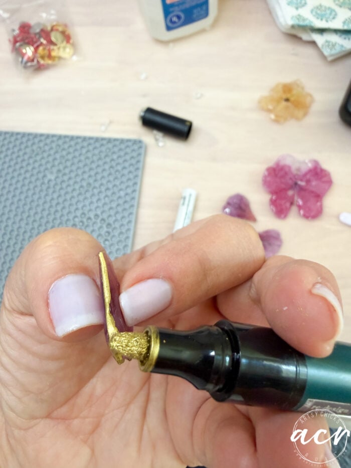 A hand with pale nail polish holds a purple resin flower while using a tool to apply gold paint to its edge&mdash;an elegant step in making DIY flower earrings; craft supplies and flowers are scattered on the table in the background.
