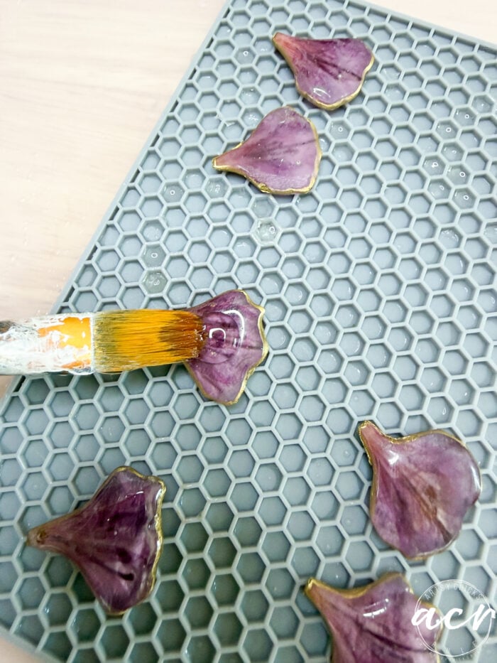 A paintbrush applies resin to purple flower petals with gold edges, arranged on a gray hexagonal-pattern silicone mat&mdash;perfect for crafting unique flower petal resin earrings or other DIY resin jewelry.