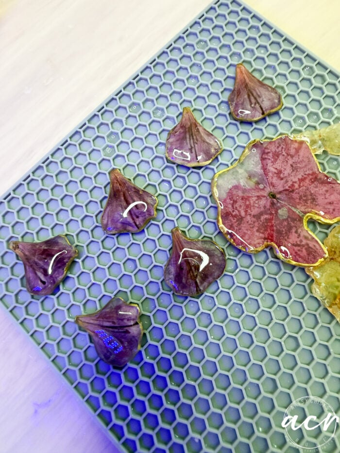 Small, glossy resin pieces shaped like purple flower petals, along with one larger pink flower-shaped piece for DIY resin jewelry, rest on a blue honeycomb-patterned mat&mdash;perfect for crafting unique flower petal resin earrings.