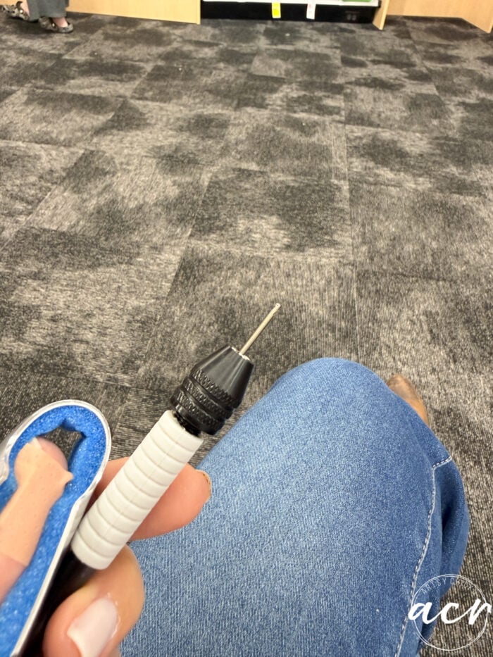 A person holding a white and black electric nail drill with a small bit attached, sitting on a gray carpeted floor with one knee bent and wearing blue jeans, prepares to work on delicate flower petal resin earrings.