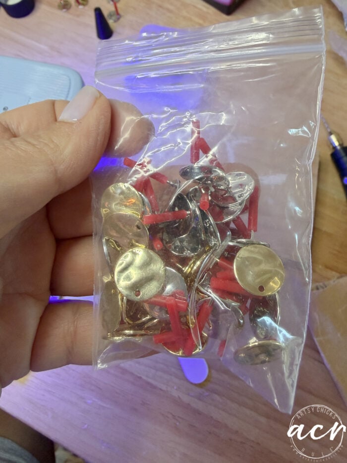A hand holds a clear plastic bag filled with gold and red metal earring findings on a wooden surface, with crafting tools in the background&mdash;perfect for creating DIY flower petal jewelry or unique flower petal resin earrings.