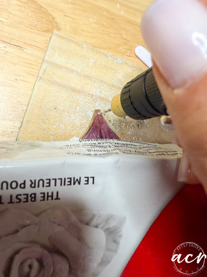 A close-up of a person using a rotary tool to drill into a purple seashell on cardboard, as part of how to make flower petal resin earrings, with newspaper partially covering the shell and wood shavings scattered around.