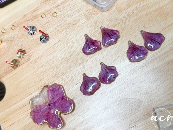 Purple and pink flower petal resin earrings, shaped like delicate petals and blooms, are arranged on a wooden surface alongside earring findings and small tools.