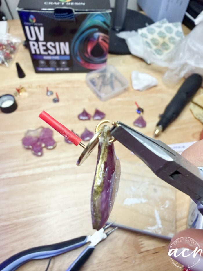 A close-up of hands using pliers to attach a metal ring to a purple resin pendant. Crafting tools, a UV resin kit, and resin earrings are scattered across the wooden workspace.