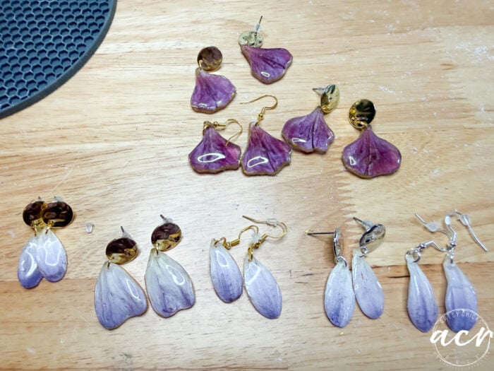 Five pairs of handmade flower petal resin earrings, crafted with real purple and white petals encased in resin, are arranged on a wooden surface near a black textured mat. The earrings feature elegant gold and silver hooks and studs.