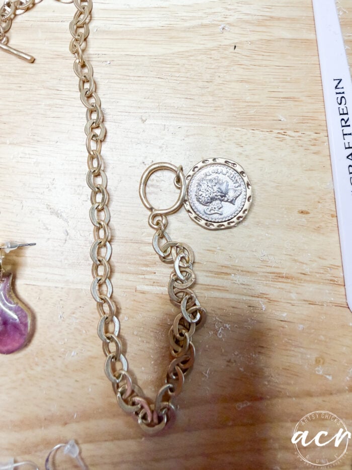 A gold chain necklace with a round coin pendant featuring a raised design is laid on a light wooden surface. Part of a purple flower petal resin earring and a white object are partially visible nearby.
