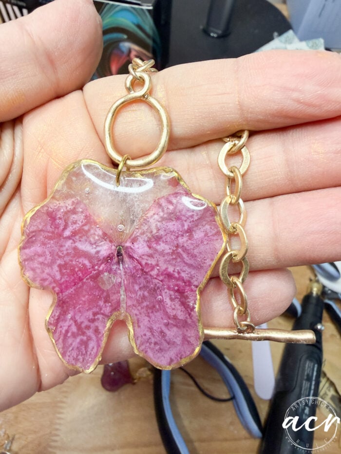 A hand holds a keychain featuring a large pink resin flower pendant with gold edges, attached to a gold chain and ring&mdash;perfect for fans of DIY resin jewelry. Jewelry-making tools are visible in the background.