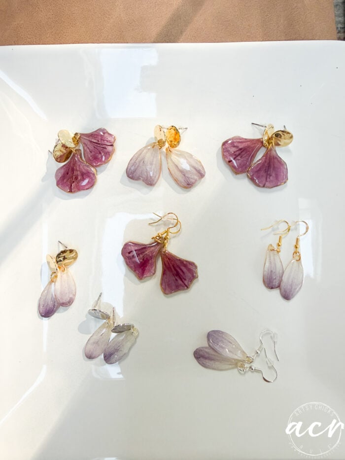 A set of five pairs of flower petal resin earrings designed to look like purple and lavender petals with gold accents, displayed on a white surface with soft natural lighting.