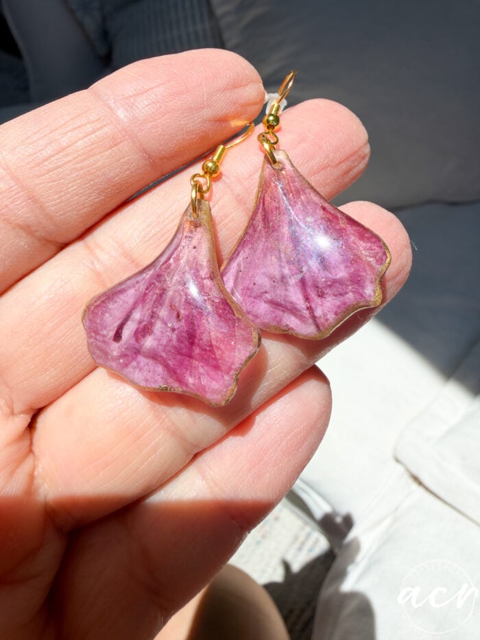 A hand holds a pair of resin earrings made from translucent pink flower petals with gold hooks, shown in bright natural sunlight.