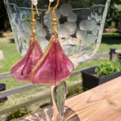 How To Make Flower Petal Resin Earrings A pair of purple, petal-shaped resin earrings with gold edges hang from a clear etched glass, set outdoors on a wooden surface with greenery in the background.
