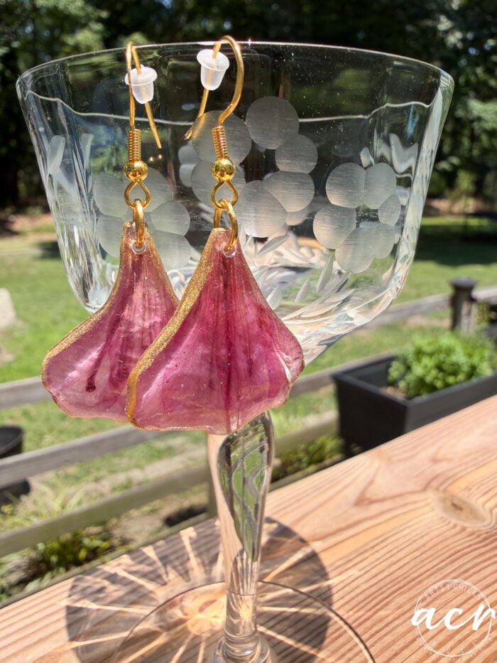 A pair of purple, petal-shaped resin earrings with gold edges hang from a clear etched glass, set outdoors on a wooden surface with greenery in the background.
