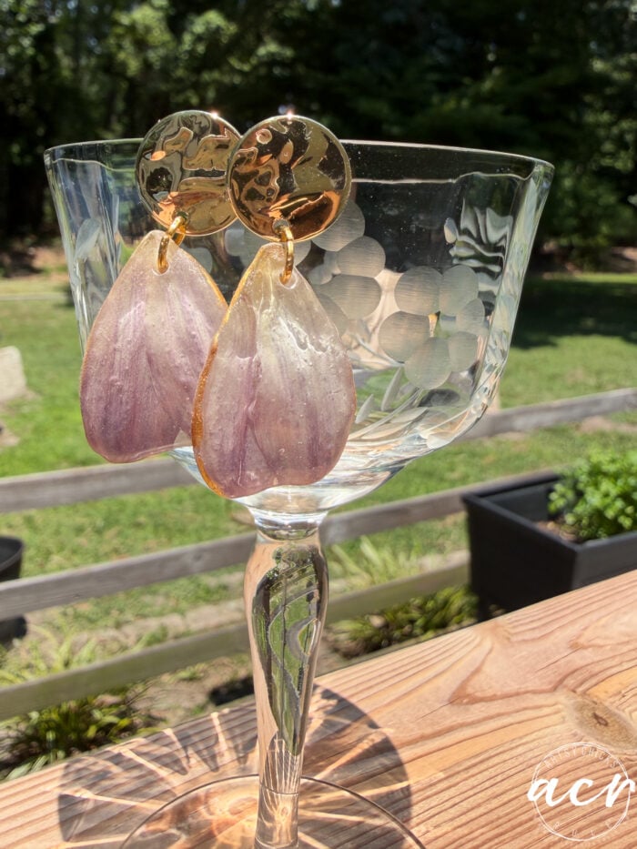 A pair of gold and lavender Flower Petal Resin Earrings hang from the rim of a vintage etched glass, set on a wooden table outdoors with greenery and garden beds in the background.
