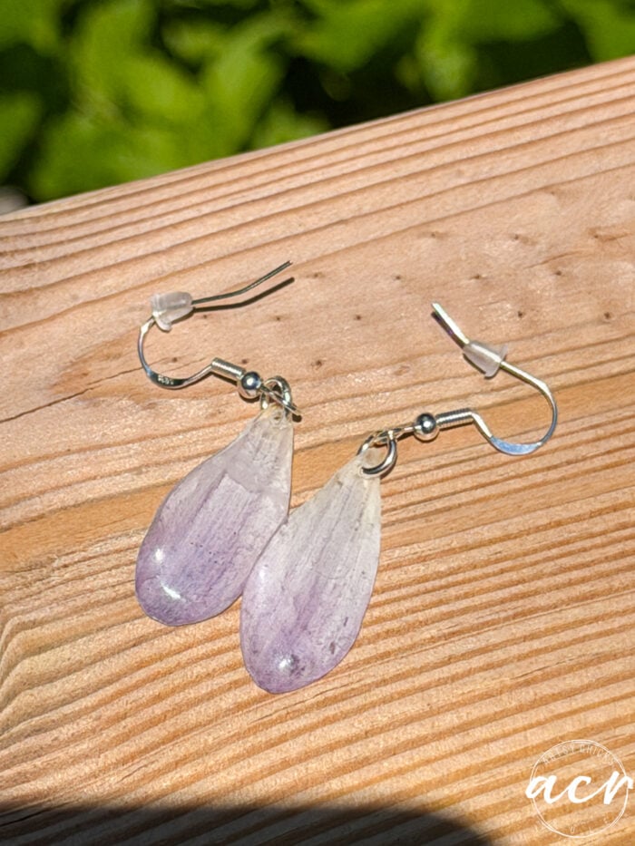 A pair of teardrop-shaped, translucent lavender resin earrings with metal hooks rest on a wooden surface. Bright sunlight highlights their delicate, glossy finish, resembling flower petal earrings. Green foliage is blurred in the background.