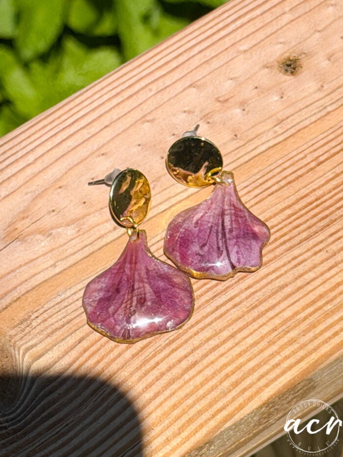 A pair of resin earrings with gold round studs and purple, petal-shaped dangles rest on a wooden surface in sunlight. A leafy green background is partially visible, with a white circular logo in the bottom right corner.