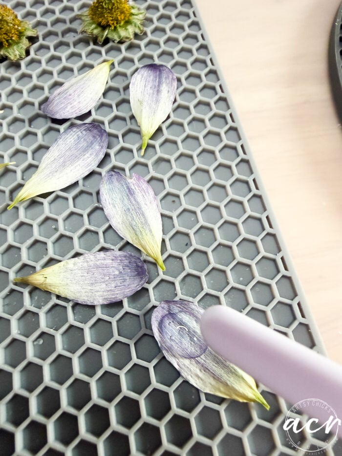 Purple and white flower petals are arranged on a gray honeycomb-patterned surface, with a hand holding a stylus pointed at one petal&mdash;perfect for creating delicate flower petal resin earrings. Some dried flowers are visible at the top.