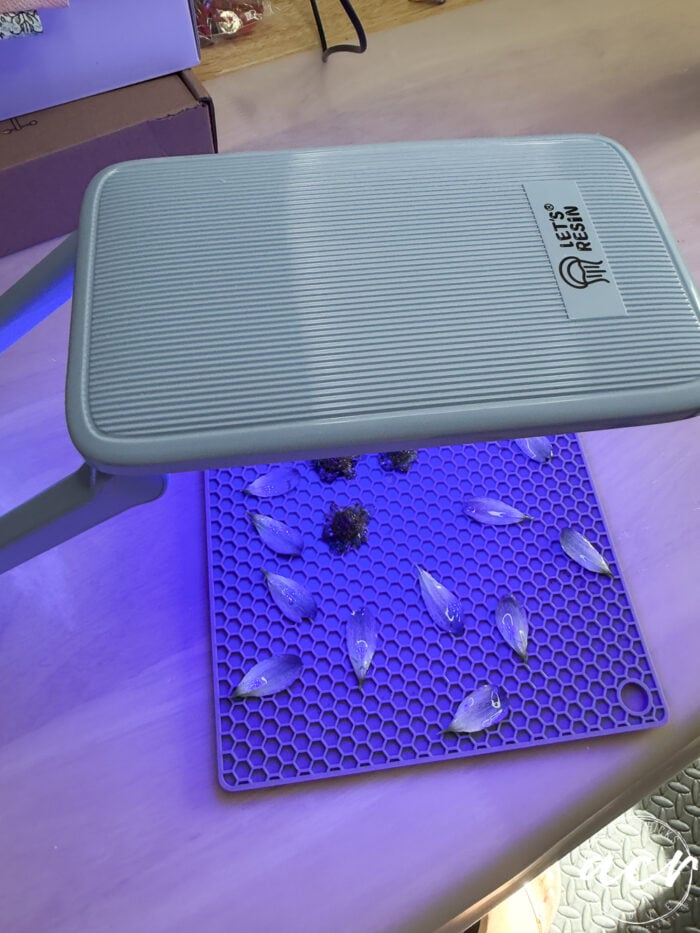 A UV lamp shines blue light onto a silicone honeycomb mat holding multiple resin-filled molds and DIY resin jewelry, with a few small pinecones on the tabletop. Nearby are a purple box and crafting supplies.
