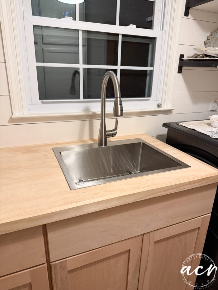 A modern kitchen sink with a stainless steel basin and faucet is set into a light wood countertop, with sleek cabinets below and a window above—perfect for both your kitchen or laundry room setup.