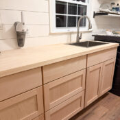 Laundry Room Update #1 (sink, cabinets & countertop) Light wood kitchen cabinets with a light wood countertop, under a window with a modern faucet and sink. White shiplap walls, open shelves with dishes, and a cordless vacuum mounted on the wall complete this fresh laundry room look.