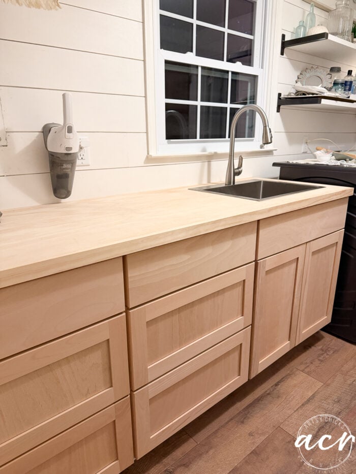 Light wood kitchen cabinets with a light wood countertop, under a window with a modern faucet and sink. White shiplap walls, open shelves with dishes, and a cordless vacuum mounted on the wall complete this fresh laundry room look.