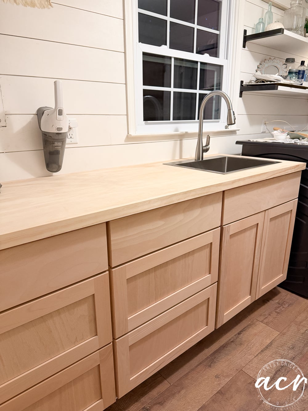 Light wood kitchen cabinets with a light wood countertop, under a window with a modern faucet and sink. White shiplap walls, open shelves with dishes, and a cordless vacuum mounted on the wall complete this fresh laundry room look.