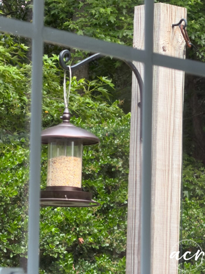 A bird feeder filled with seeds hangs from a metal hook on a wooden post, seen through window grid panes in the laundry room. Lush green bushes and trees create a vibrant backdrop beyond the cabinets and sink.