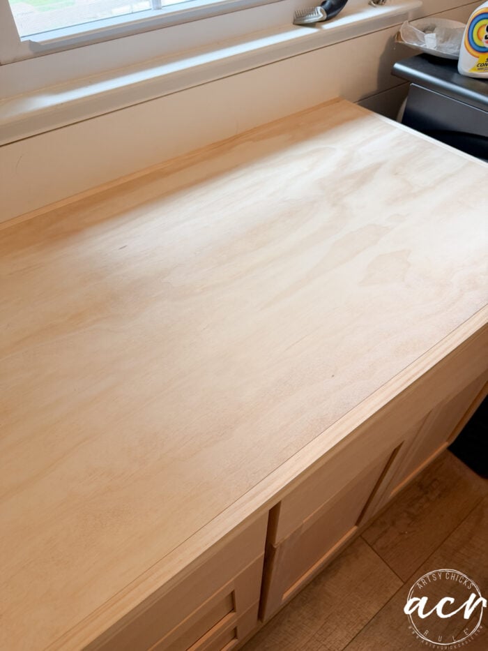 A light wood countertop with a clean, unfinished plywood surface sits above light wood cabinets and a sink, next to a window and a stove in the kitchen.