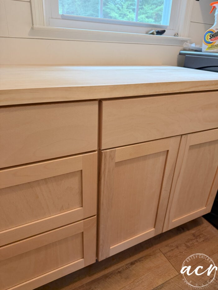 Light wood kitchen cabinets with shaker-style doors and drawers sit beneath a white countertop and sink. A window with white trim and cleaning supplies in the background enhance this laundry room. The floor is wooden.