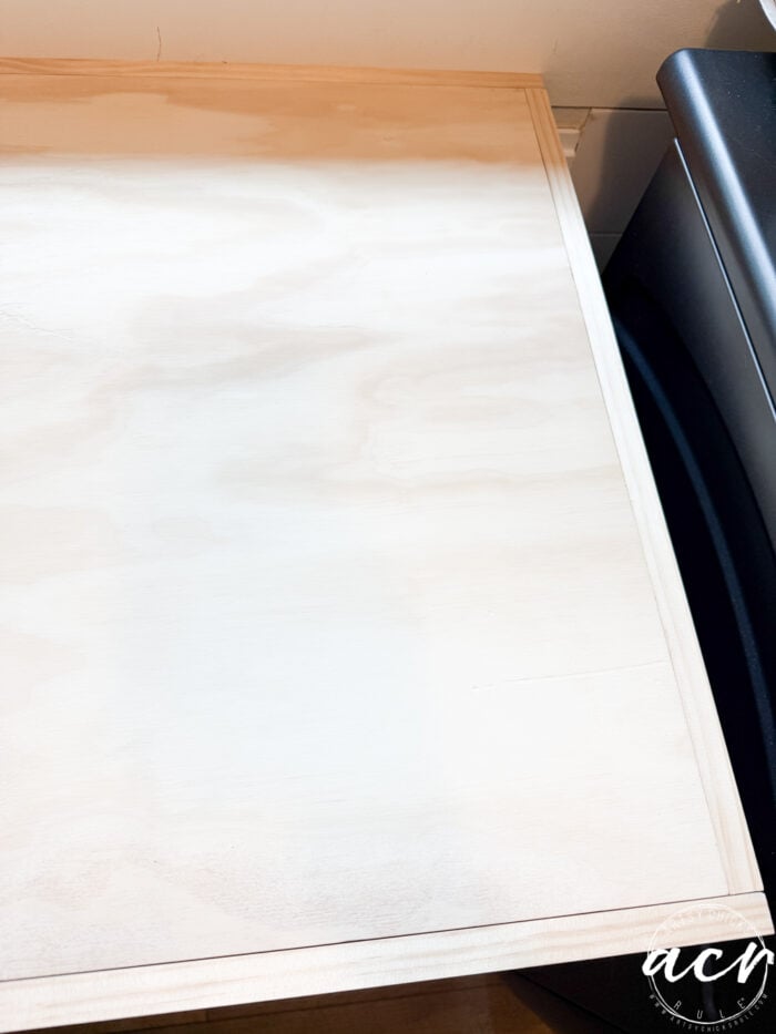 A sheet of light-colored plywood with a rectangular trim border rests on top of a black appliance, next to a wall—ideal for cabinets or as a sink countertop in a laundry room. A small watermark with the letters acn appears in the lower right corner.