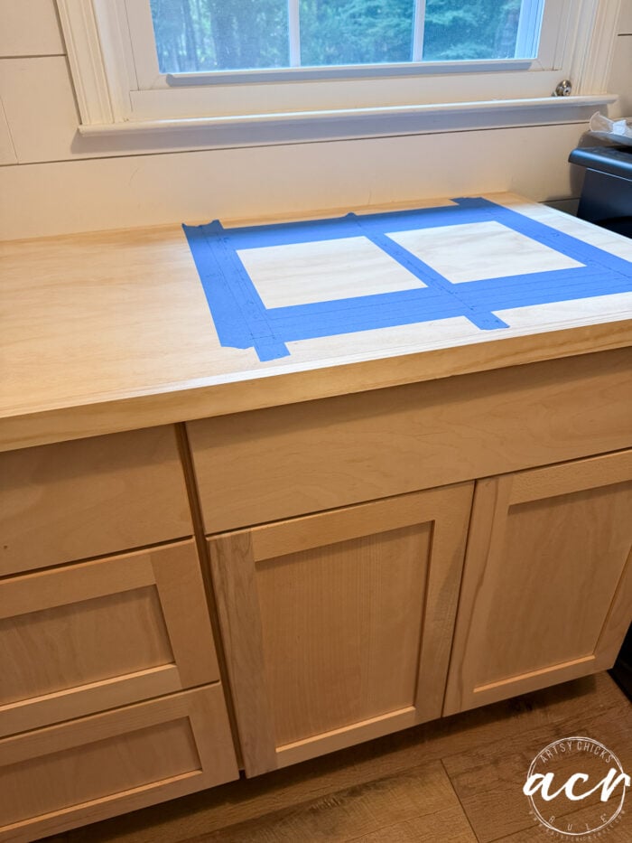 A wooden kitchen counter with blue painter’s tape outlining two rectangular sections sits below a window. Light wood cabinets and drawers, perfect for storage, are underneath the counter.