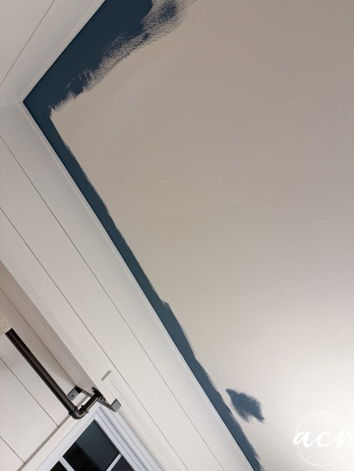 A ceiling partially painted with blue along the edges, leaving the center unpainted. The photo, taken from an angle in a laundry room, also shows part of a white wall, window, curtain rod, and upper cabinets.