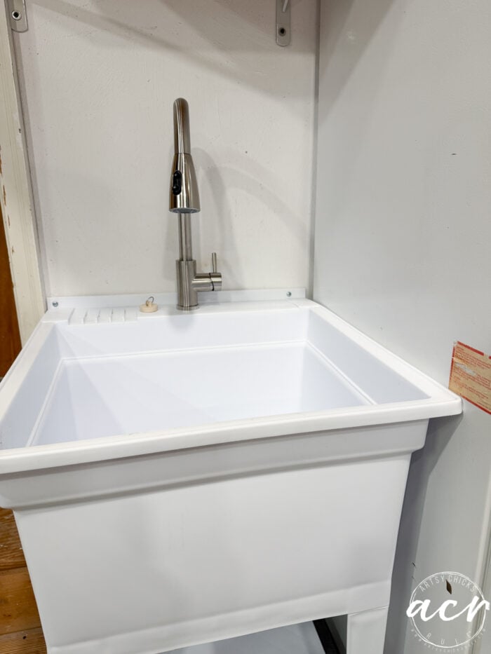 A white utility sink with a modern stainless steel faucet is installed against a white wall in a small laundry room. The sink sits on sturdy legs and has a small drainboard area on the left side, perfect for everyday laundry tasks.