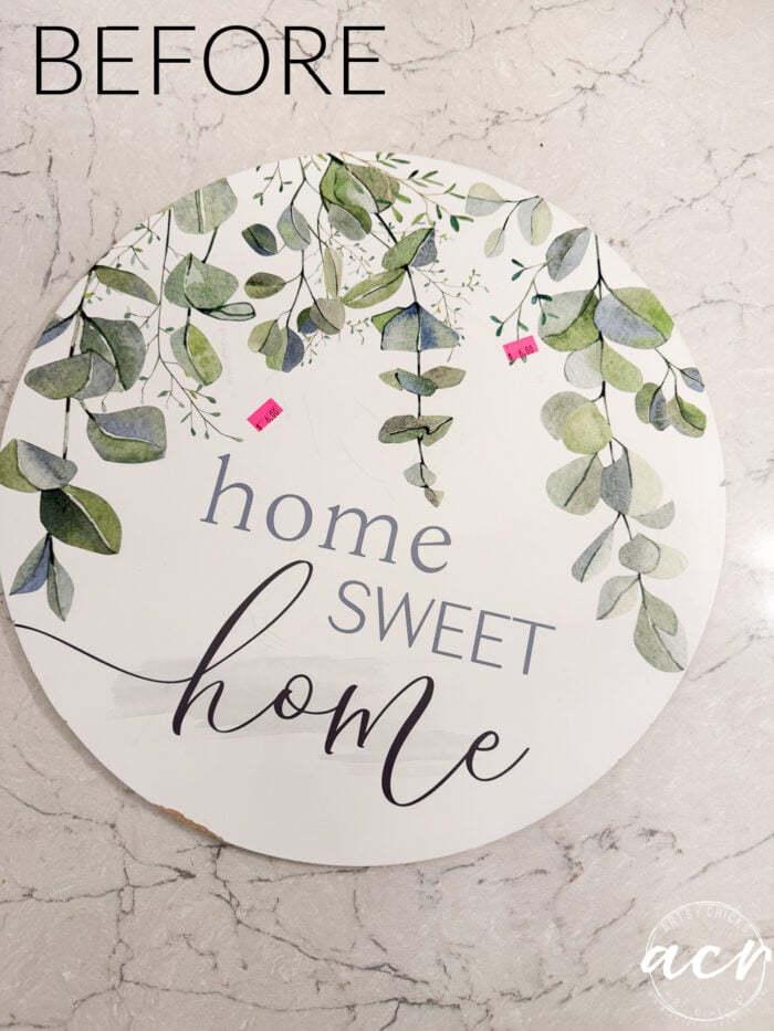Round sign on a marble surface with green leafy design. Text reads home sweet home. Two small pink price tags are visible on the front. The word BEFORE appears at the top left, highlighting this charming Fall Door Hanger perfect for welcoming guests.