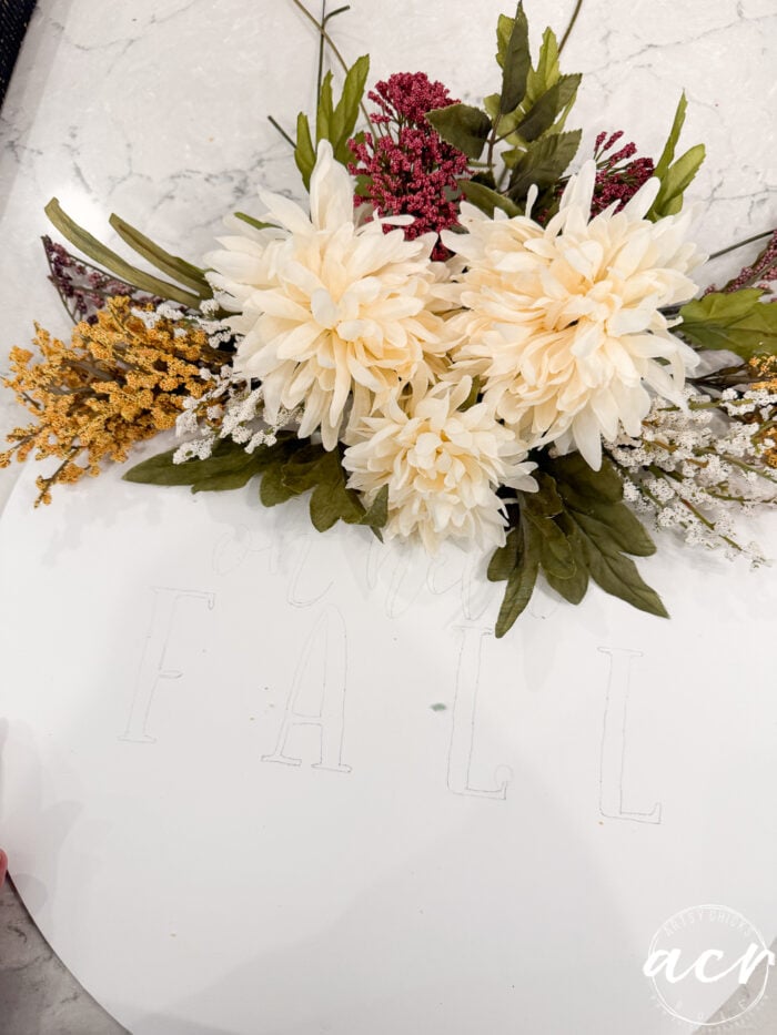 A floral arrangement of artificial cream, yellow, and purple flowers sits above lightly outlined letters spelling FALL on a white surface, creating a charming Fall Door Hanger to welcome the season.