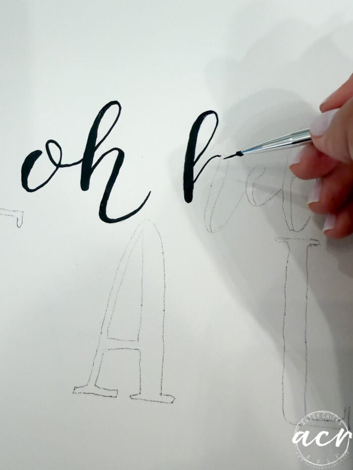 A hand uses a small brush and black ink to fill in cursive handwritten letters of “h” on white paper, with faint pencil outlines of other letters visible—perfect inspiration for creating personalized Hello Fall Decor or a charming Fall Door Hanger.